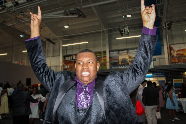 A student wearing a gray suit with purple accessories smiles and makes the rock and roll symbol with his hands  