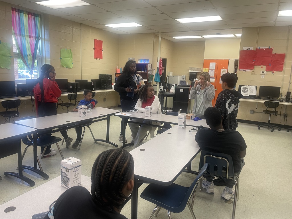 An indoor classroom setting where a group of middle-school-aged students and an instructor are gathered around tables practicing clinical skills. A woman in a grey hoodie stands at the head of a table, demonstrating the use of a stethoscope. Several students are seated or standing nearby, some with blood pressure cuffs wrapped around their arms, while boxes of medical equipment sit on the tables. The room has a computer lab setup along the back wall, beige cinderblock walls, and colorful paper decorations near the windows.