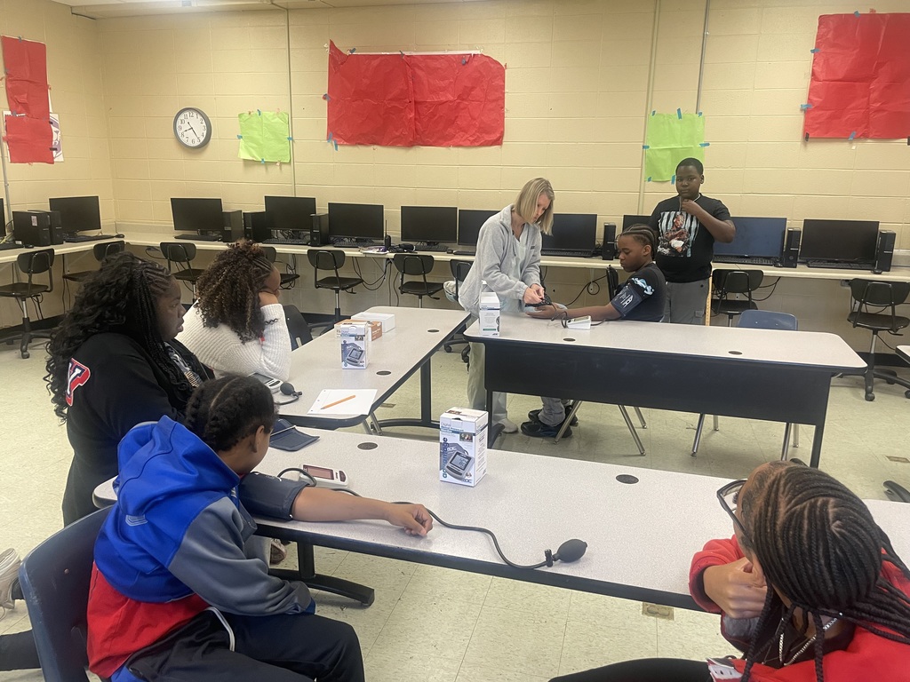 A classroom scene where an instructor and students are practicing how to measure blood pressure. In the center, a female instructor in a grey zip-up hoodie leans over a table to assist a seated student with a blood pressure cuff. Other students sit at surrounding tables, observing the demonstration or looking at digital monitors. In the foreground, a student in a blue and red jacket has a cuff attached to their arm. The background shows a row of desktop computers along a beige wall decorated with red and green paper.