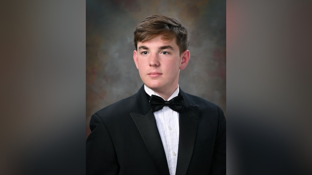 Jaxon Oden, senior at Corner High School looks ahead and poses for a photo in front of a blue and brown background. He wears a black tuxedo with a white shirt underneath it.
