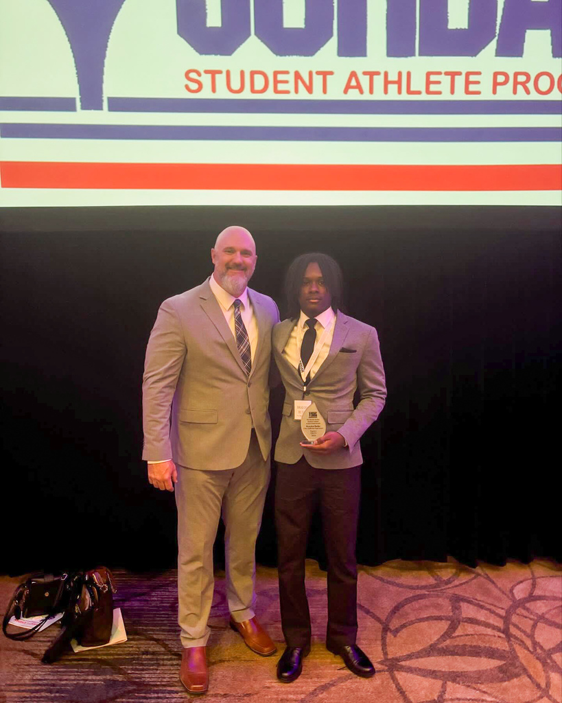 Clay-Chalkville High School student Brayden Butler and football coach Stuart Floyd stand together for a photo inside a ballroom. They both wear suits. Brayden holds a Bryant-Jordan Student-Athlete Achievement Award in his hand.