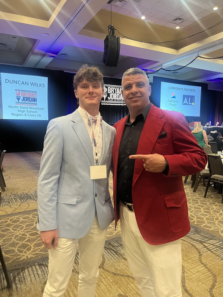Oak Grove High School student George Dorman poses for a photo with Chris Musso in a ballroom. George wears a blue blazer with tan pants and Chris wears a red blazer with tan pants. Chris points to George in the photo.