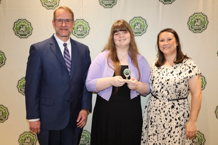 Corner High School’s Emily McDaniel stands between two adults in front of a backdrop with the Alabama Farm-City Committee logo on it. The man wears a suit and Emily and the other woman wear dresses. Emily holds an award in her hand.