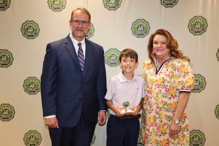 A man, woman and child pose in front of a photo backdrop. The man wears a suit and the woman wears a dress The boy wears a collared shirt with dress pants. The boy smiles and holds a trophy.
