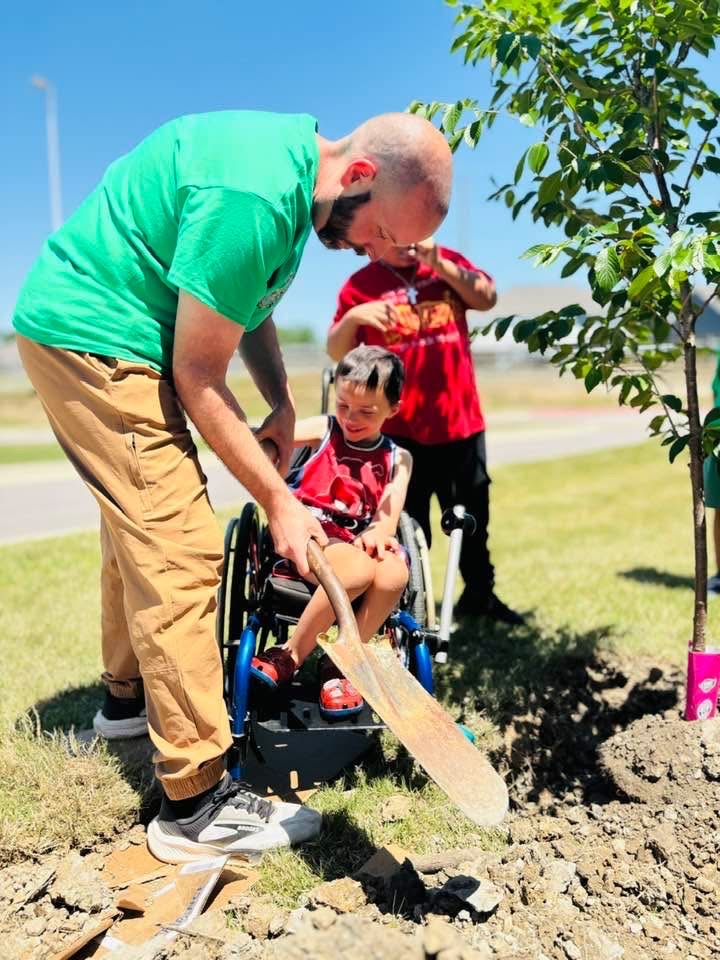 A man helps a student who is in a wheelchair dig a hole for a tree outside a school.