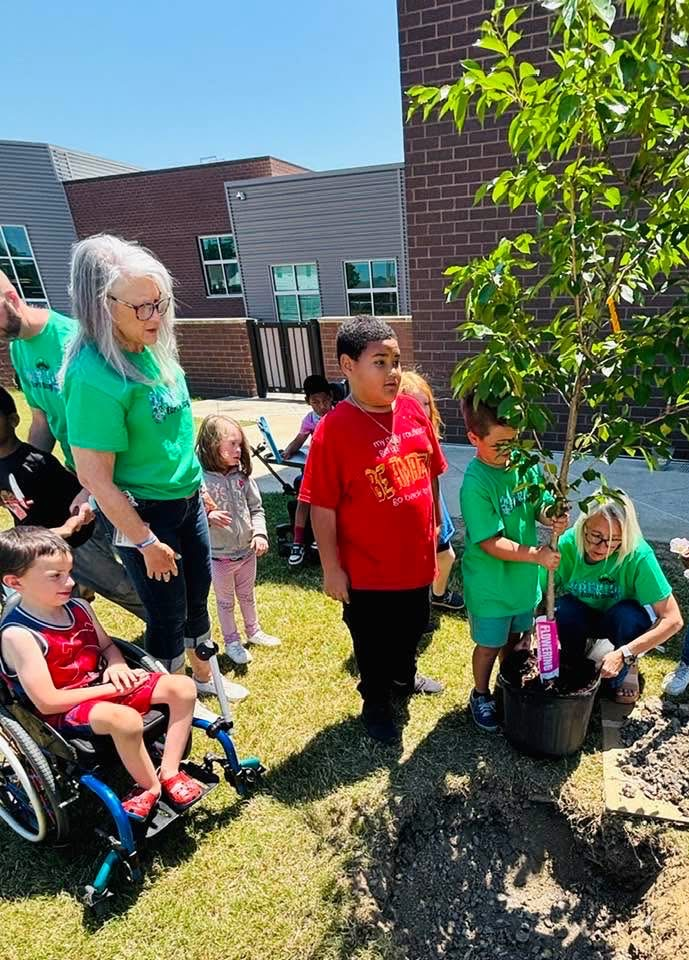 A classroom of students stand and sit outside a school and watch one of the students plant a tree.