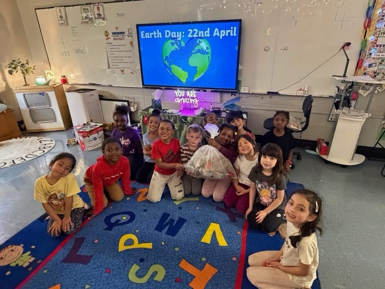 A group of students sit on the floor of a classroom and smile while holding a bag of trash that they collected from outside.