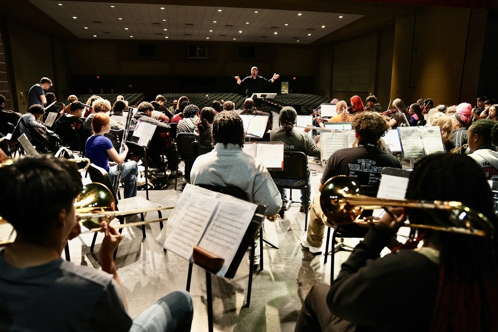 A director leads a band while they sit in chairs on a stage and play instruments.
