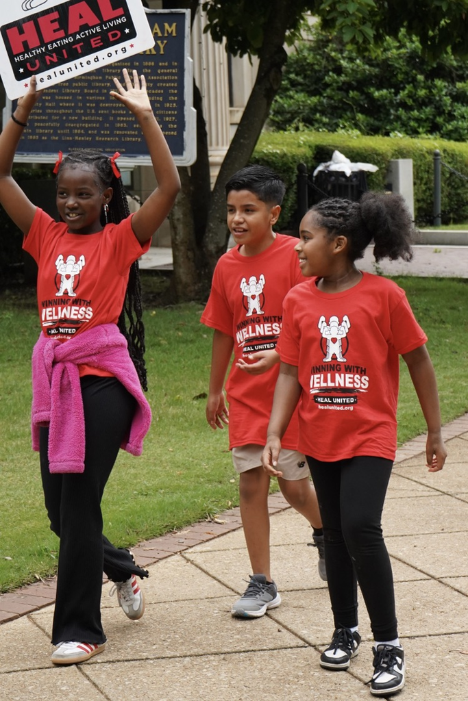 Students smile as they walk down a sidewalk. They wear matching “Healthy Eating Active Living” (HEAL) shirts and one student holds up a HEAL sign.