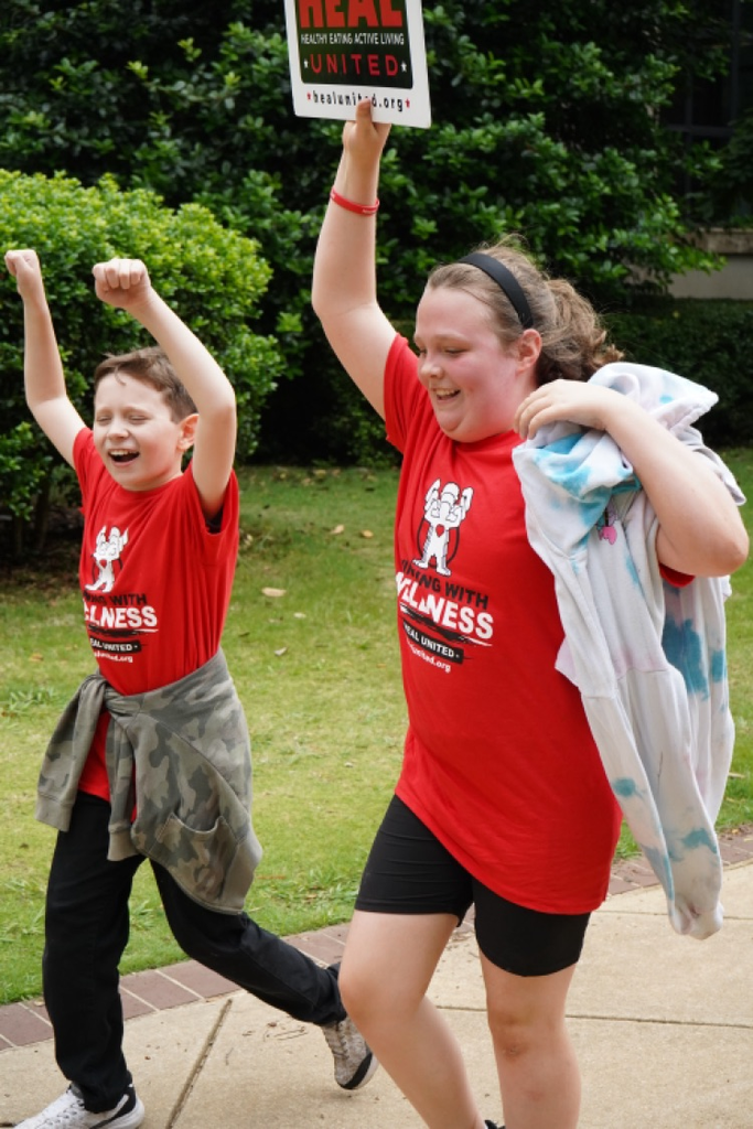Students walk down a sidewalk smiling with their hands in the air. They wear matching “Healthy Eating Active Living” shirts.
