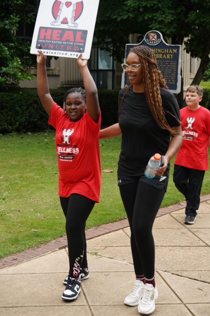 A student and an adult smile while walking down a sidewalk. The student holds a “Healthy Eating Active Living” sign.