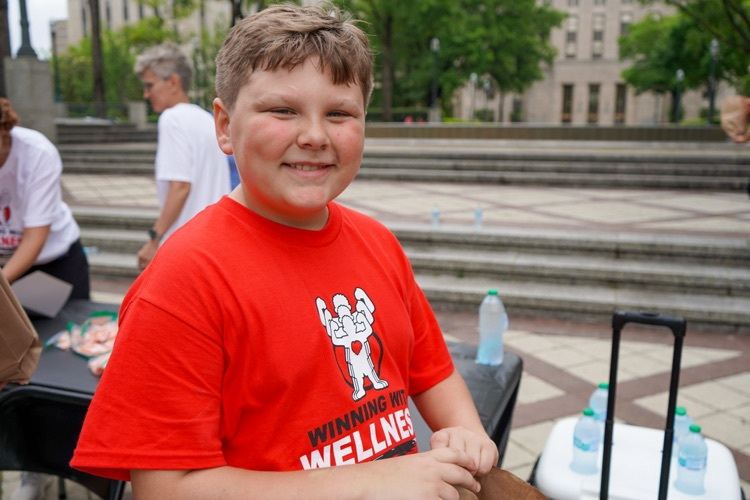 A boy smiles while standing at a park. He wears a “Healthy Eating Active Living” shirt.