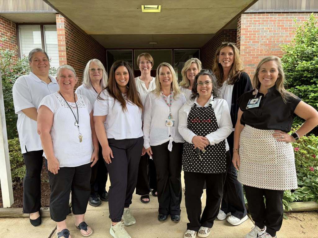A group of women stand together for a photo and smile. Some of the women wear white shirts and black pants. Three of the women wear aprons over their clothes.