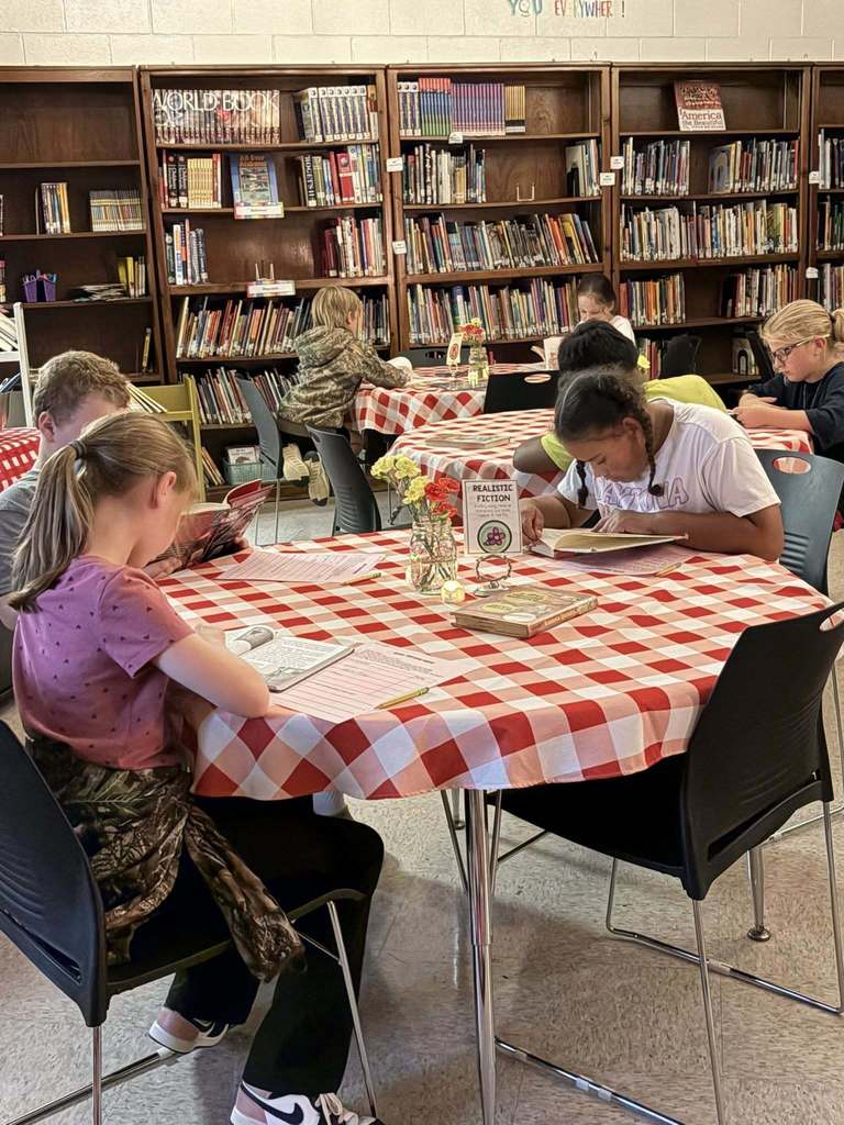 Students sit at a table with a red checkered table cloth and read books inside a library.