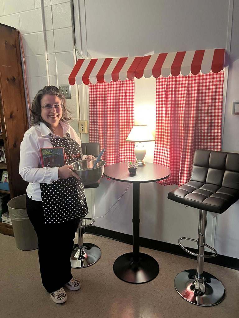 A woman wears an apron and stands in a room decorated like a cafe. She holds a bowl and smiles.