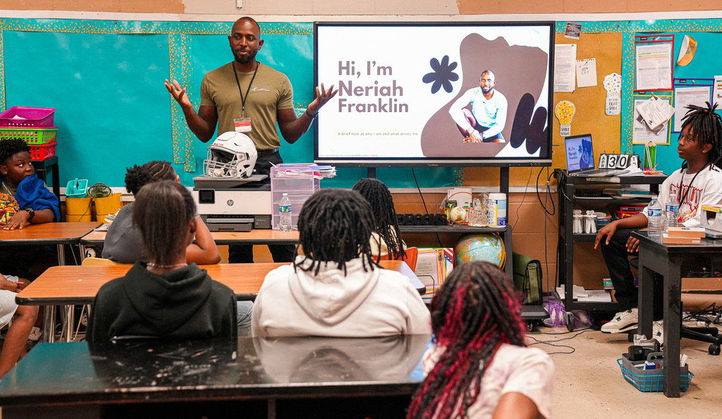 A man gives a presentation to a classroom of students. The screen behind him says "Hi, I'm Neriah Franklin" and a football helmet sits in front of him.