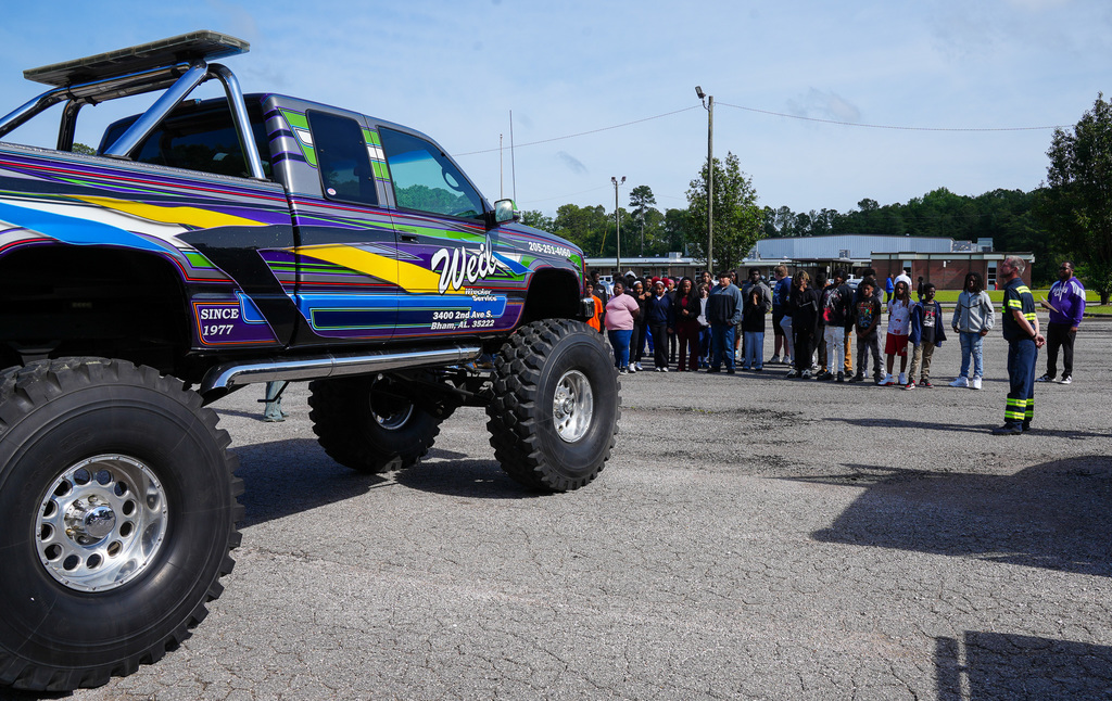 Students stand in a parking lot and look at a monster truck that says "Weil Wrecker Services."