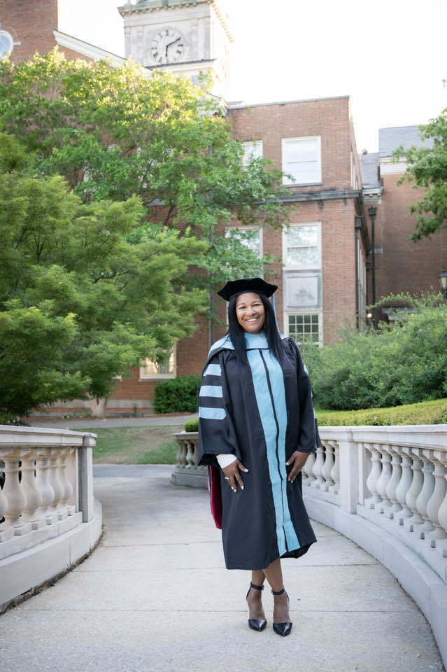 Center Point Elementary School assistant principal Dr. Theta Warren stands outside a building on a walkway and smiles. She wears a graduation cap and gown.
