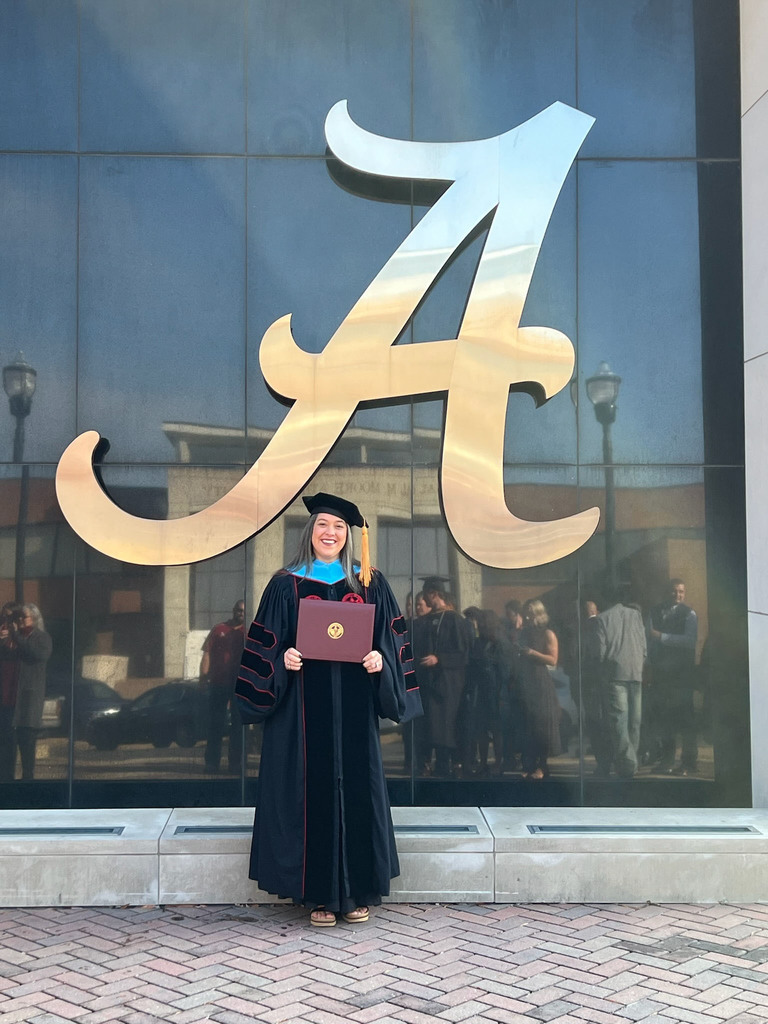 Bryan Elementary School principal Dr. Brandi Wilson stands in front of a building with the Alabama script A on the side of it. She wears a graduation cap and gown and holds a diploma in her hand.