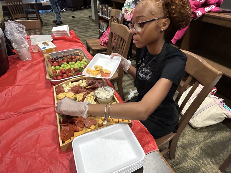 A student sits at a table and puts crackers and meat onto a plate.
