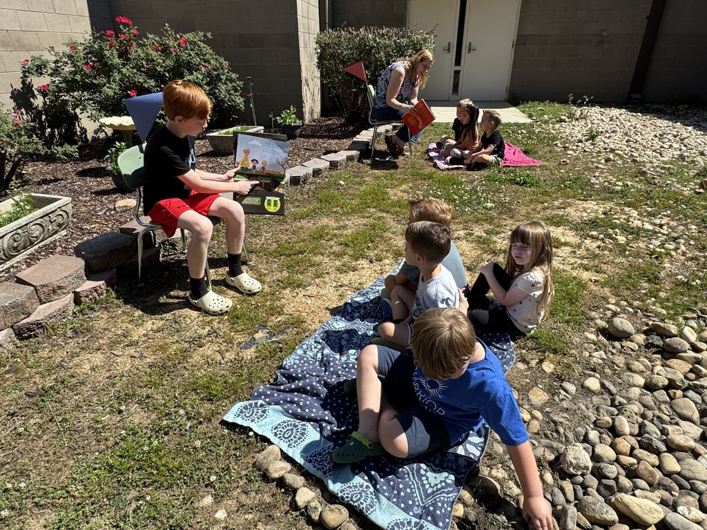 A fourth grade boy reads a garden book to Pre-K students who are sitting on a blanket outside.
