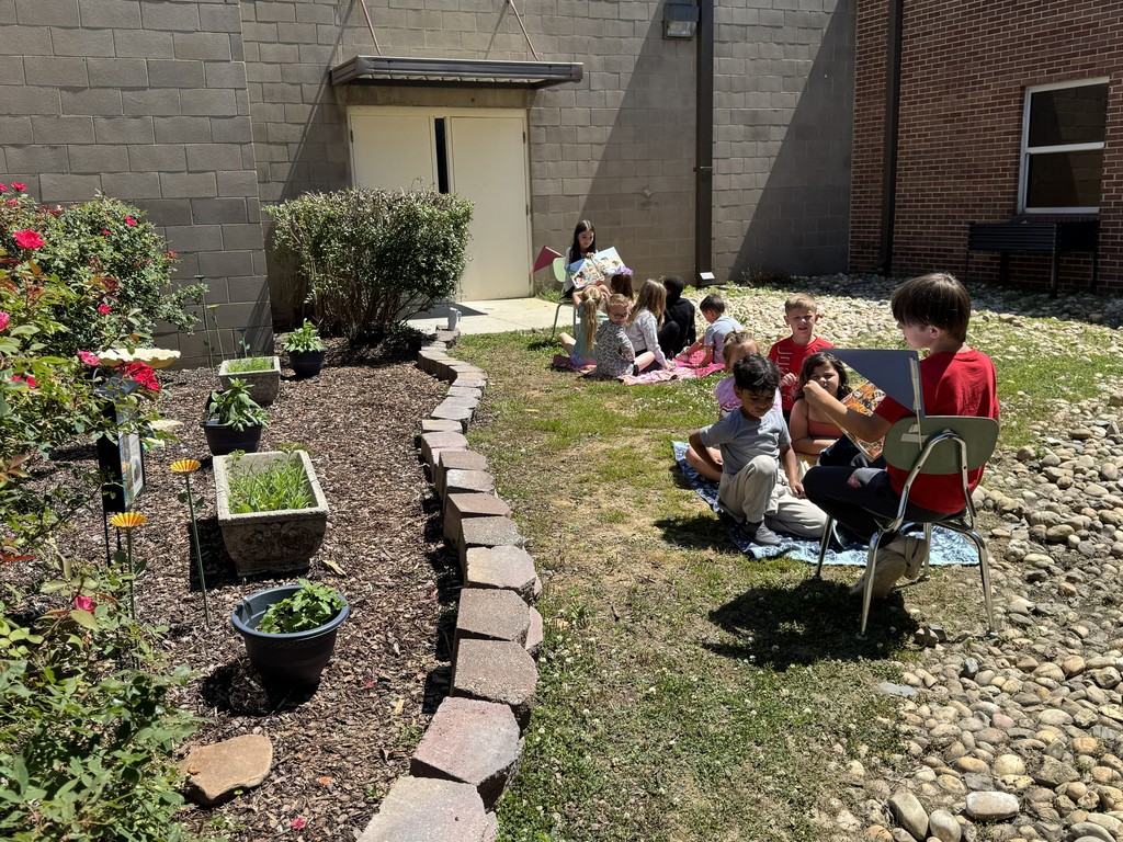 Fourth grade students read a garden book to Pre-K students who are sitting on blankets outside.