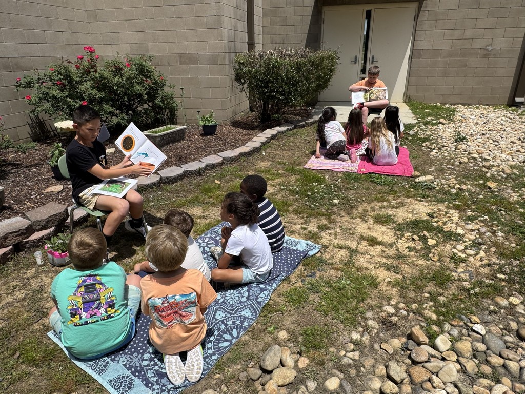 A fourth grade boy reads a garden book to Pre-K students who are sitting on a blanket outside.