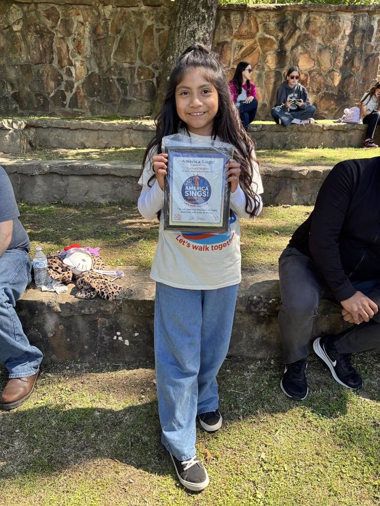 A student stands outside and smiles as she holds an America Sings plaque in her hands.