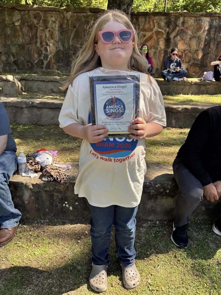 A student stands outside and smiles for a photo. She holds an America Sings plaque in her hands.