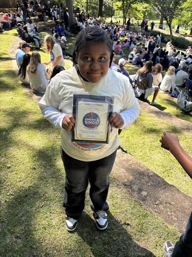 A student stands outdoors and smiles for a photo. She holds a plaque that says "America Sings".