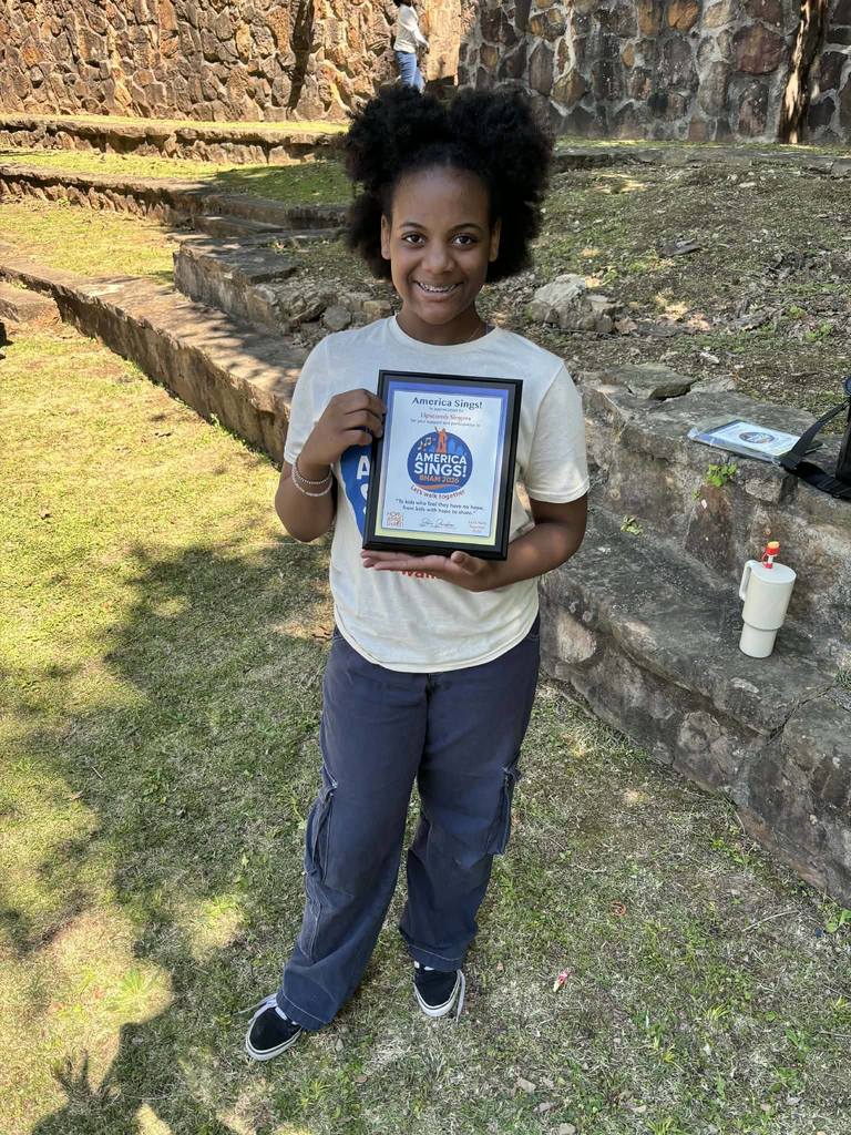 A girl smiles while standing outside. She holds a plaque that says "America Sings".
