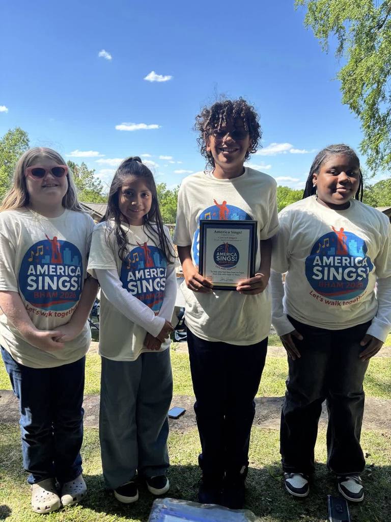A group of students stand together for a photo outdoors. Each of the students wear shirts that say "America Sings" on them. One of the students holds a plaque in his hand.