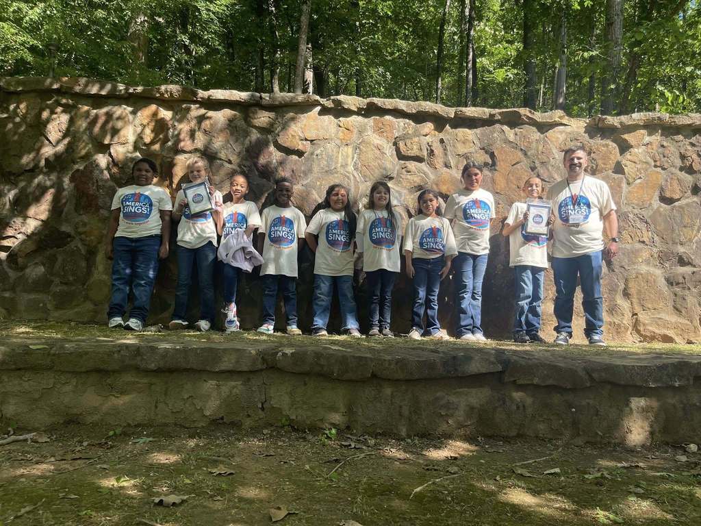 A group of students stand together outdoors for a photo with their choir teacher. They smile. Two of the students hold plaques in their hands.