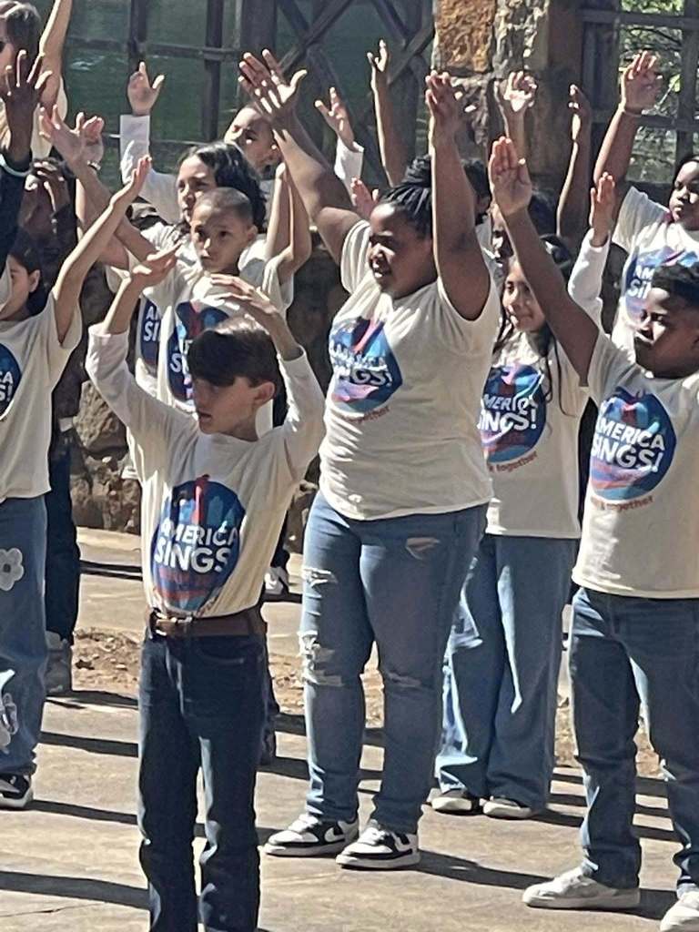 Students wearing matching "America Sings" shirts hold their arms up as they perform a song outdoors.