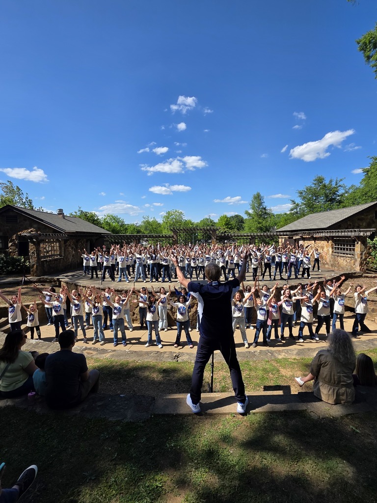 A choir director leads a large choir of students. They perform at an outdoor amphitheater.