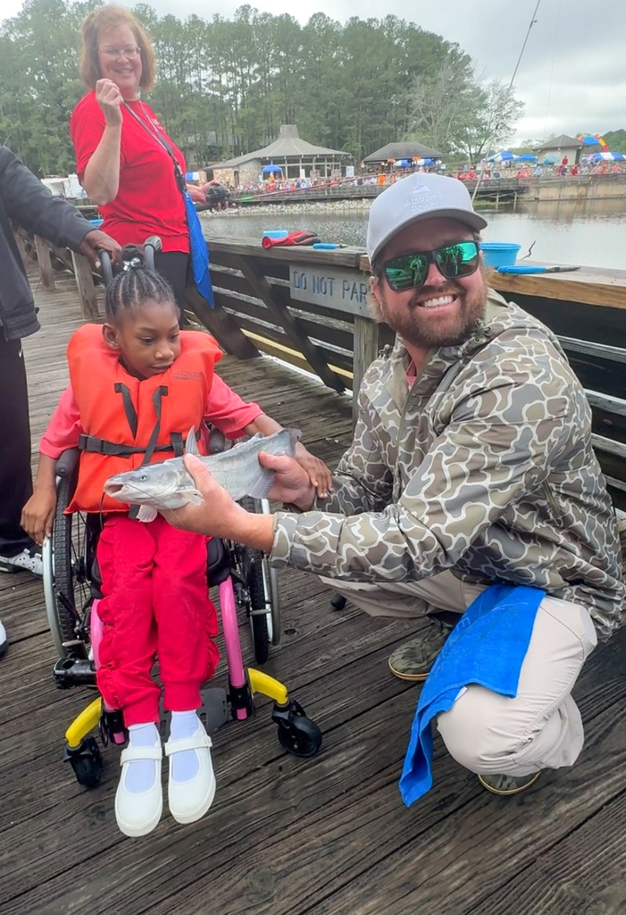 A man and a girl stand and sit on a dock. The man smiles as he holds a catfish out toward the girl. The girl looks at the fish and touches the man's arm.