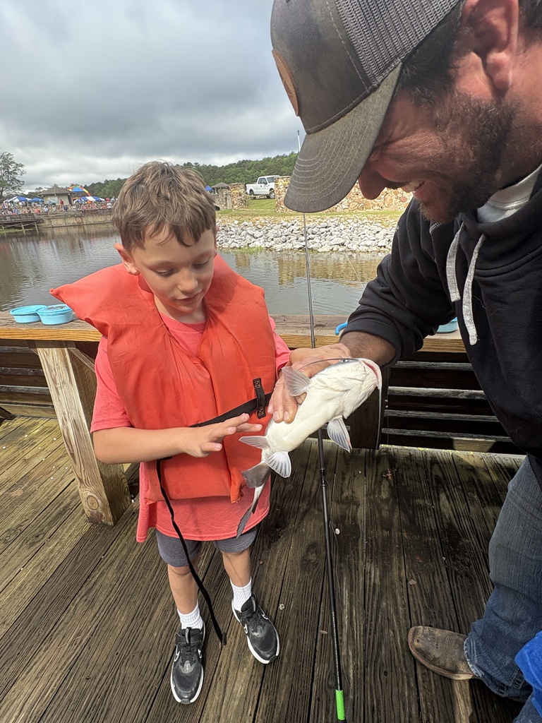 A man and a boy stand on a doc. The man holds out a catfish for the boy to touch.