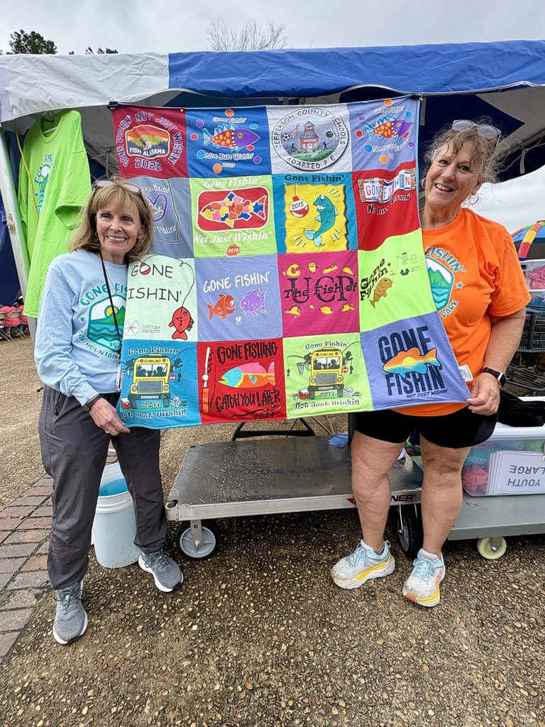 Two women stand together outside for a photo. They both hold up the ends of a quilt with t-shirt designs on it.