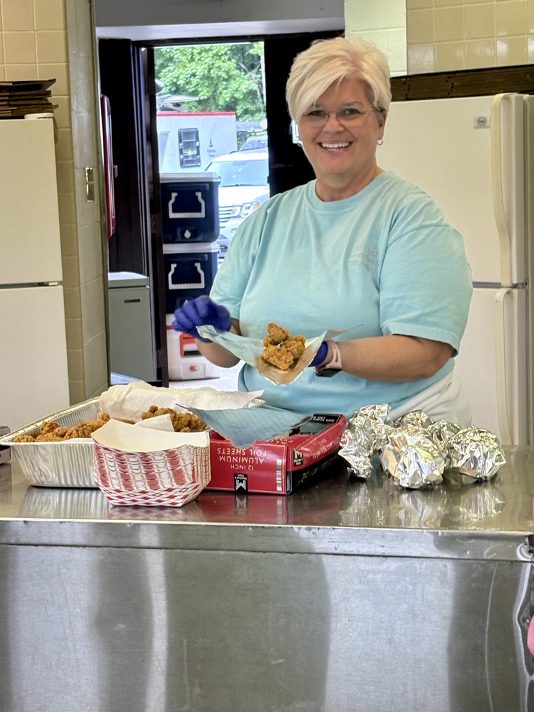 A woman smiles while she puts fried catfish into aluminum foil. She stands in a kitchen while she works.