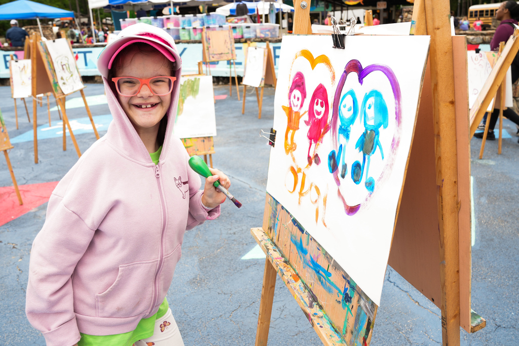 A girl stands outside and paints. She looks at the camera and smiles while holding a paint brush in her hand.