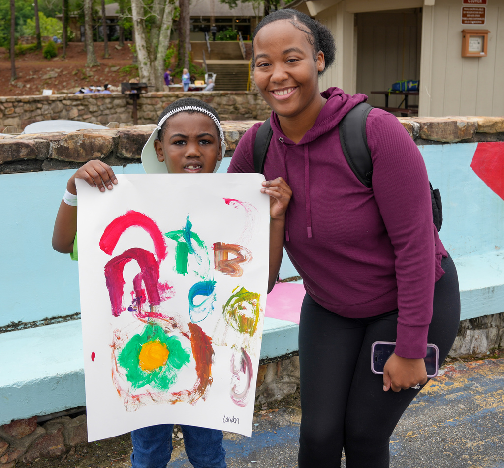 A woman and a boy stand together and smile for a photo. The boy holds up a piece of paper with a painting on it.