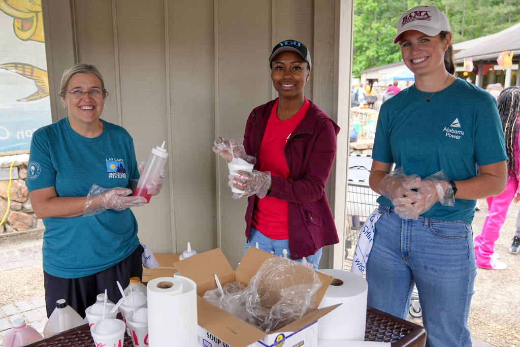 Three woman stand together at a picnic table outdoors. They hold snow cone ingredients and smile.