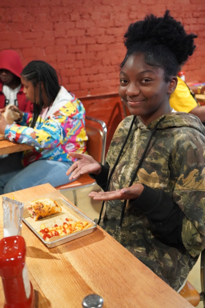 A student sits at a restaurant table and smiles. She points her hands toward her meal.