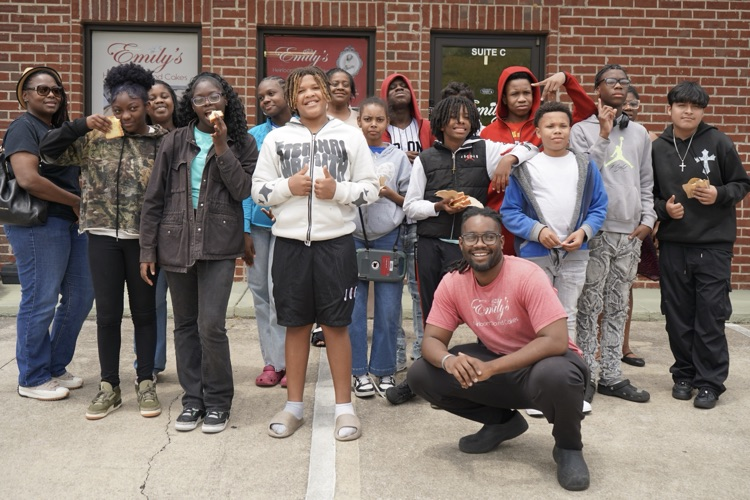 Students and adults stand together and smile for a photo outside a business.