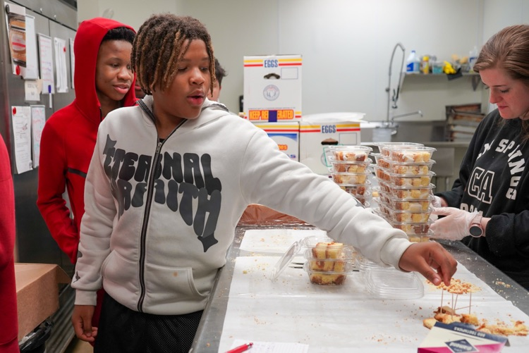 A boy reaches for a piece of pound cake on a table inside a bakery kitchen.