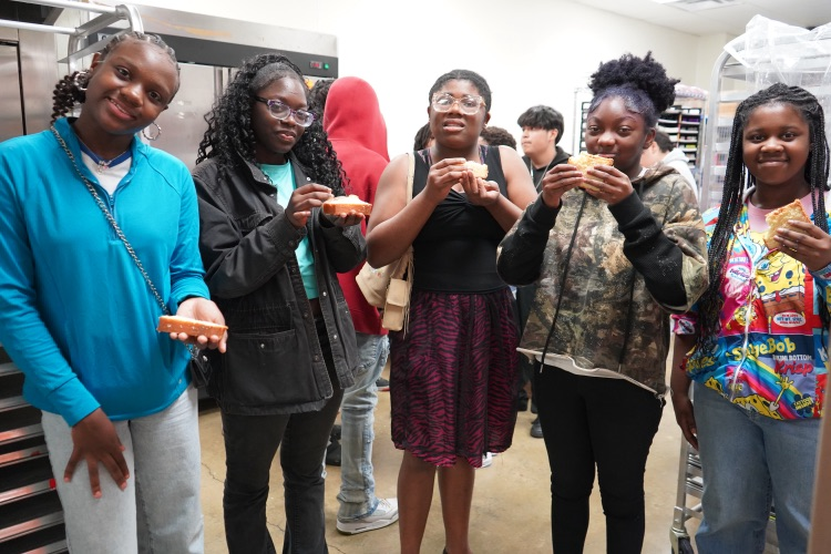 Students smile and hold up pieces of pound cake while standing inside a kitchen of a bakery.