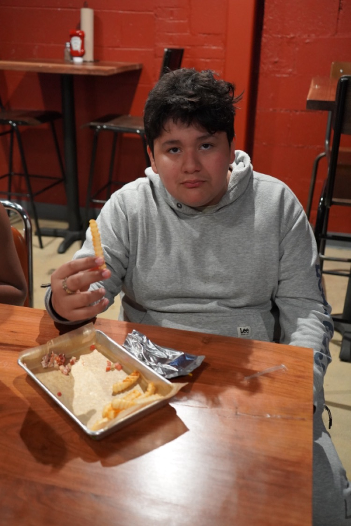 A student sits at a table at a restaurant and eats a fry.