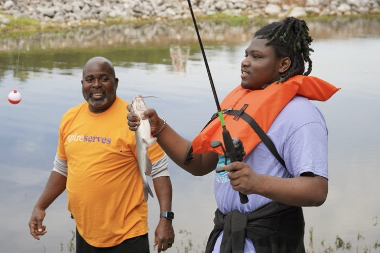 A man and a boy stand by a lake. The boy holds a fishing rod and a fish in his hands.