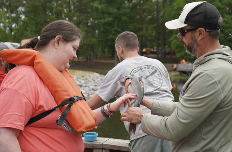 A man and a girl stand on a fishing pier. The man holds a catfish out toward the girl. 