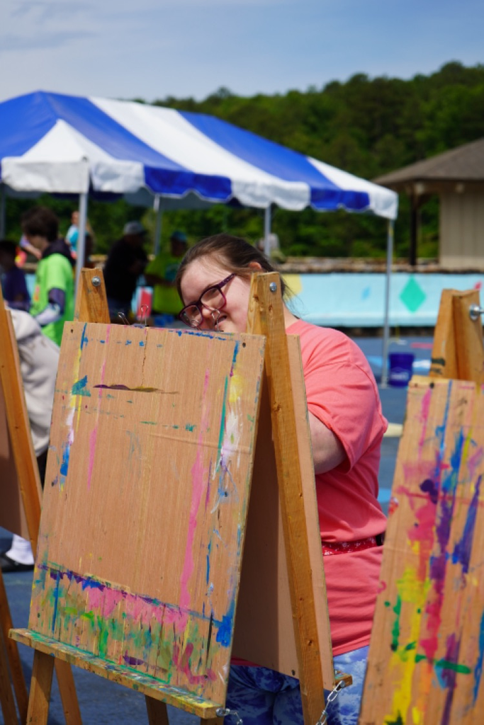 A girl smiles while painting a piece of paper on an easel outdoors.